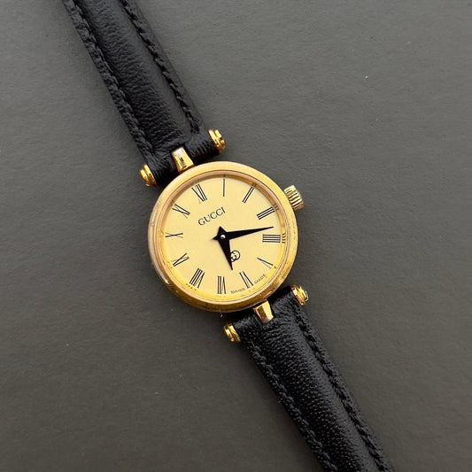Gucci Sherry gold-tone women's watch with black leather strap on a dark background.