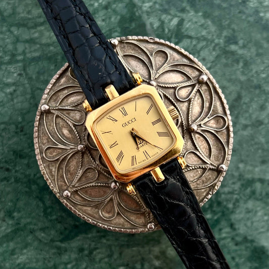 Gucci Sherry gold soft-square watch with black croc-style strap displayed on a metal stand.