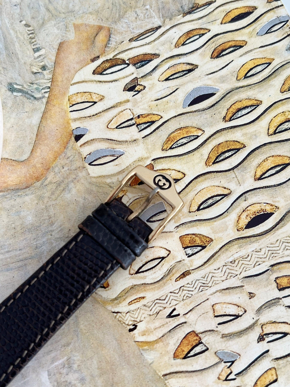 Close view of the Gucci Sherry women’s watch gold tone GG pin buckle, black leather keepers and textured strap.
