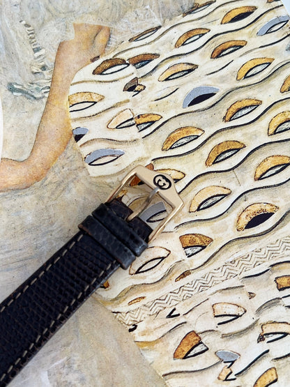 Close view of the Gucci Sherry women’s watch gold tone GG pin buckle, black leather keepers and textured strap.