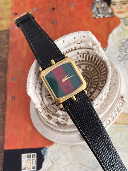 Gucci Sherry women’s watch with a green and red dial, square gold tone case and black textured leather strap, styled on a Colosseum ornament.