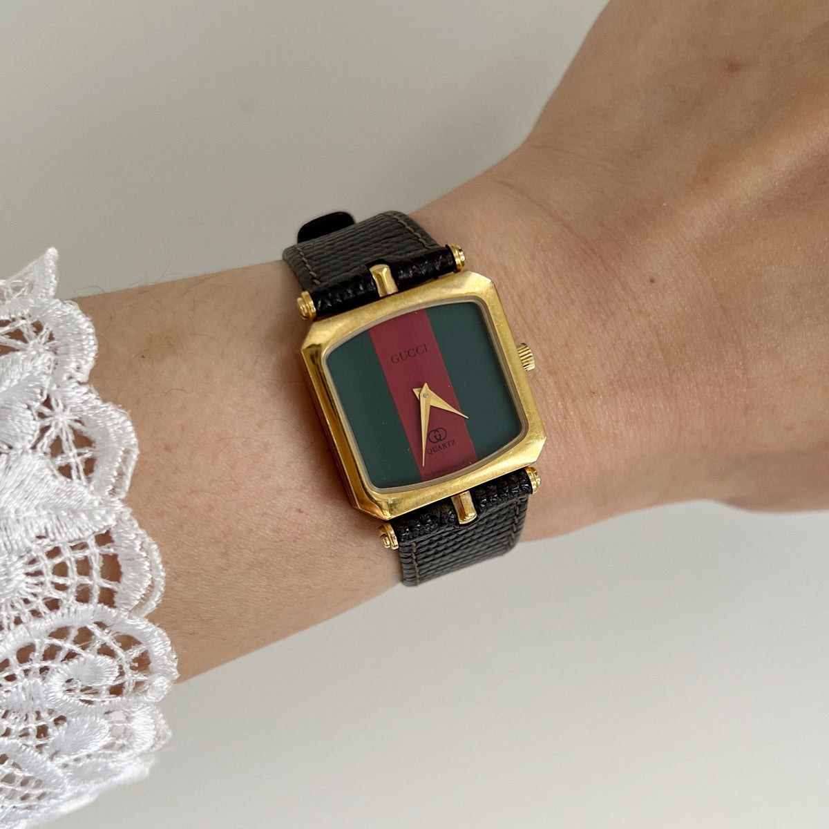 Gucci Sherry women’s watch on the wrist, with a green and red stripe dial, square gold tone case and black textured leather strap.