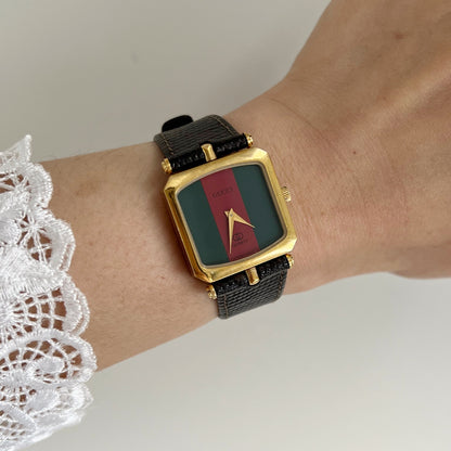 Gucci Sherry women’s watch on the wrist, with a green and red stripe dial, square gold tone case and black textured leather strap.