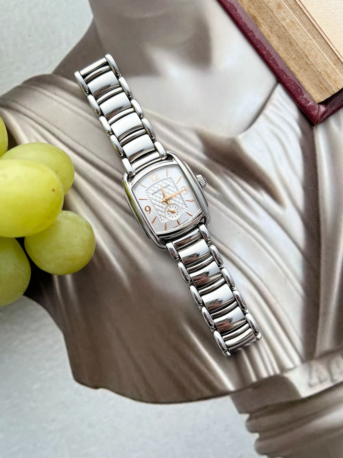 Hamilton Bagley bracelet watch laid across a sculpted silver backdrop, showing its curved rectangular case and polished steel links.