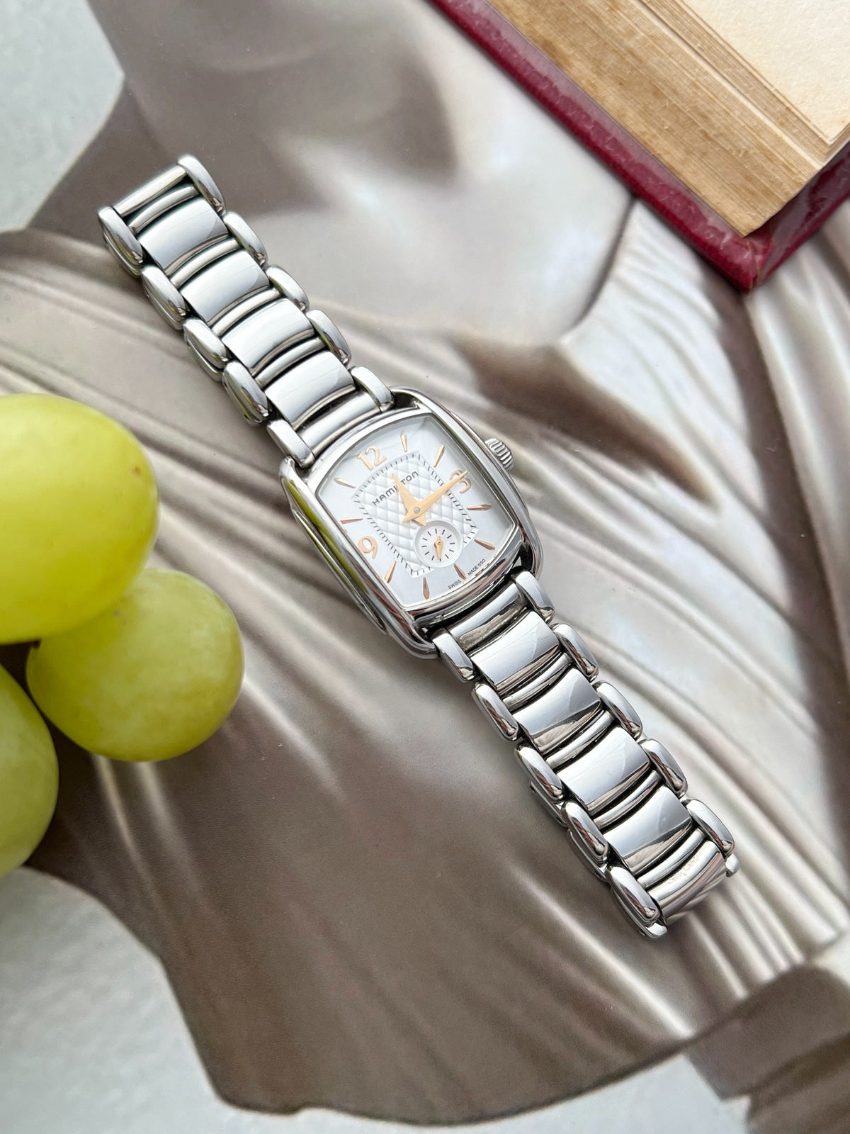 Hamilton American Classic Bagley watch with a silver quilted dial, rose gold tone hands and polished steel bracelet beside grapes and an open book.