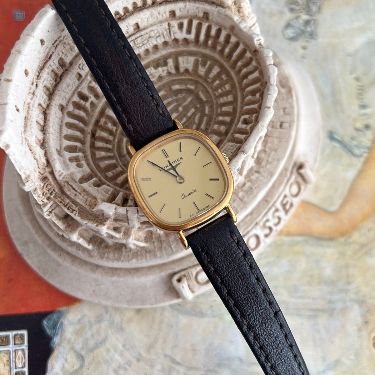 Longines women’s quartz watch with a champagne baton dial, gold tone cushion case and original black leather strap beside a Colosseum model