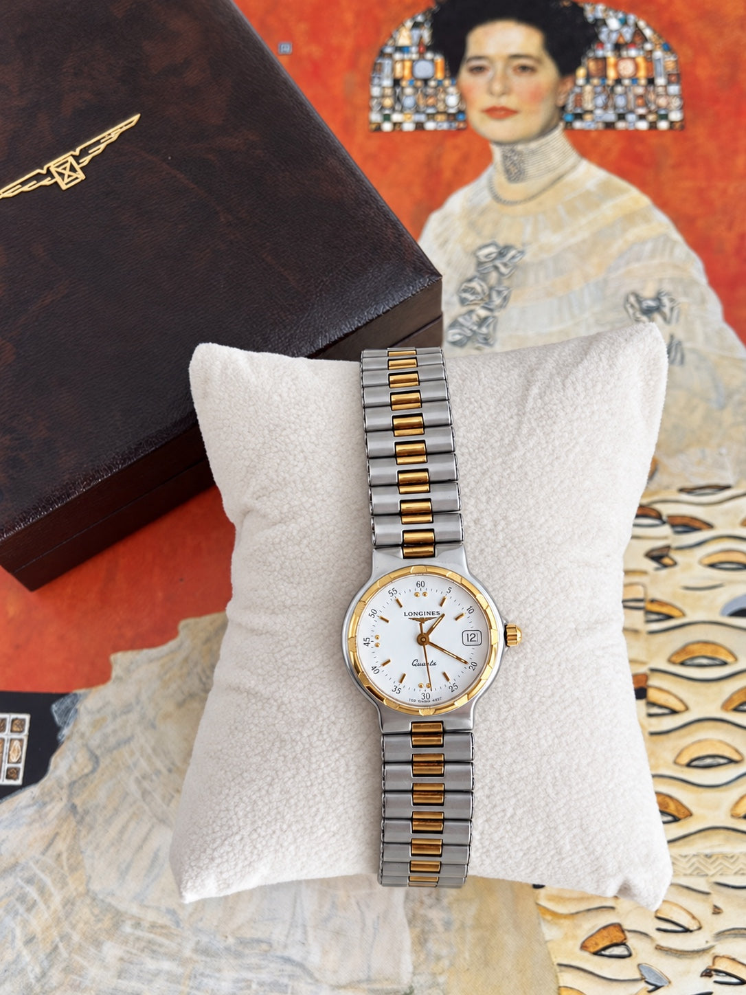 Longines Conquest women's watch with a round silver tone case, gold tone bezel, white dial and two tone link bracelet on a cushion beside a Longines box.