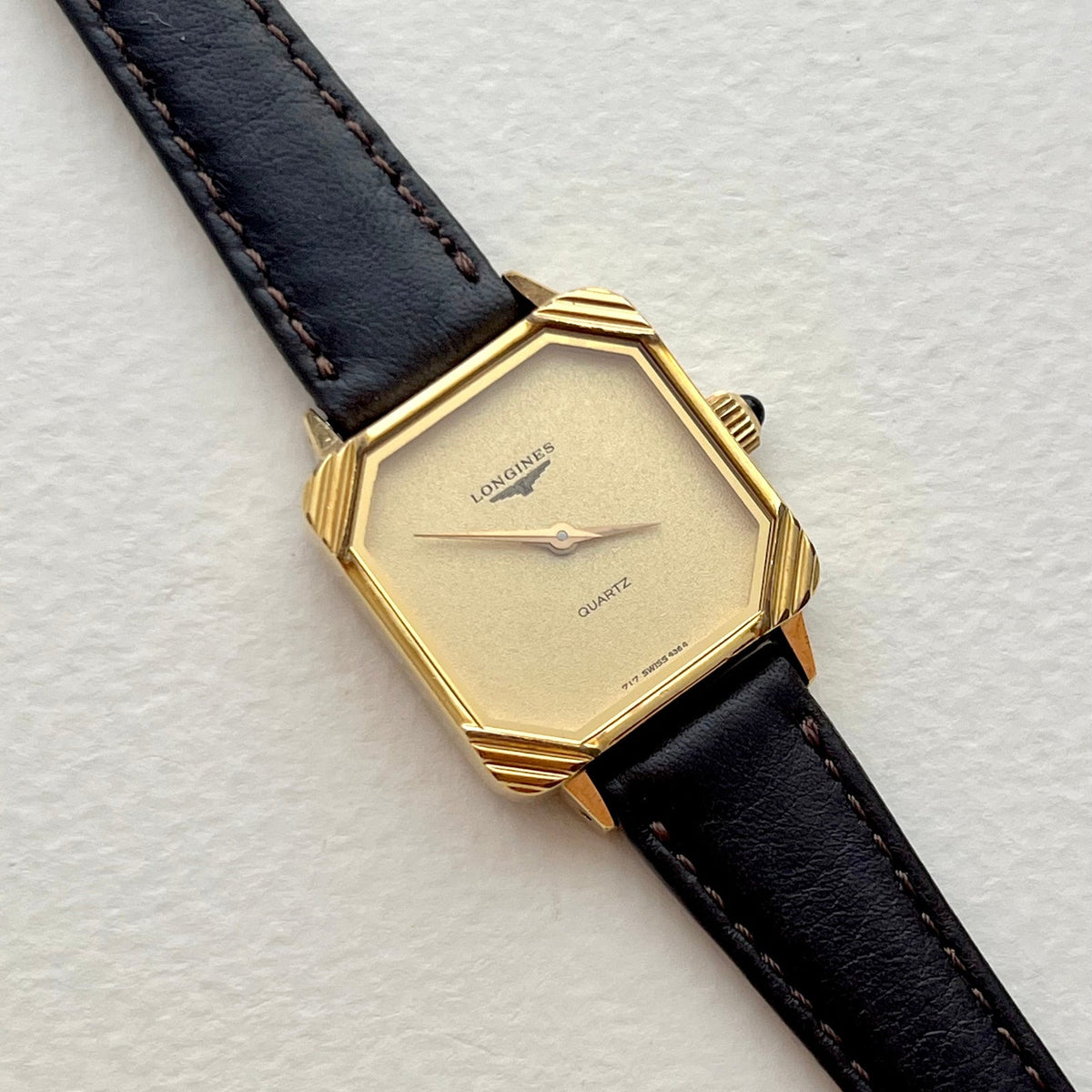 Longines ladies watch with black leather strap, minimalist champagne dial and sculpted gold corner case, on a light textured background.
