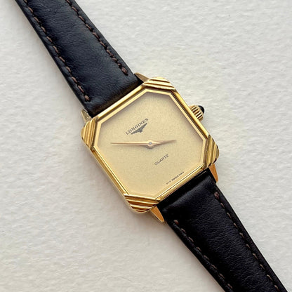 Longines ladies watch with black leather strap, minimalist champagne dial and sculpted gold corner case, on a light textured background.
