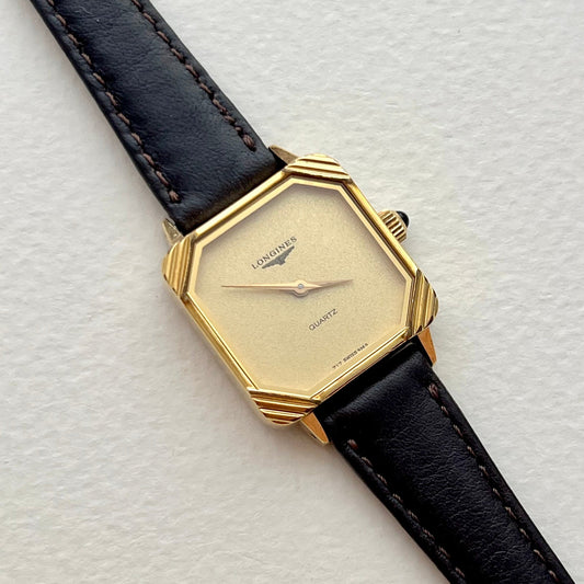 Longines ladies watch with black leather strap, minimalist champagne dial and sculpted gold corner case, on a light textured background.