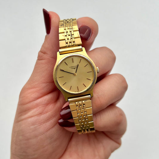 Longines round gold manual-wind watch held in hand against a plain background.