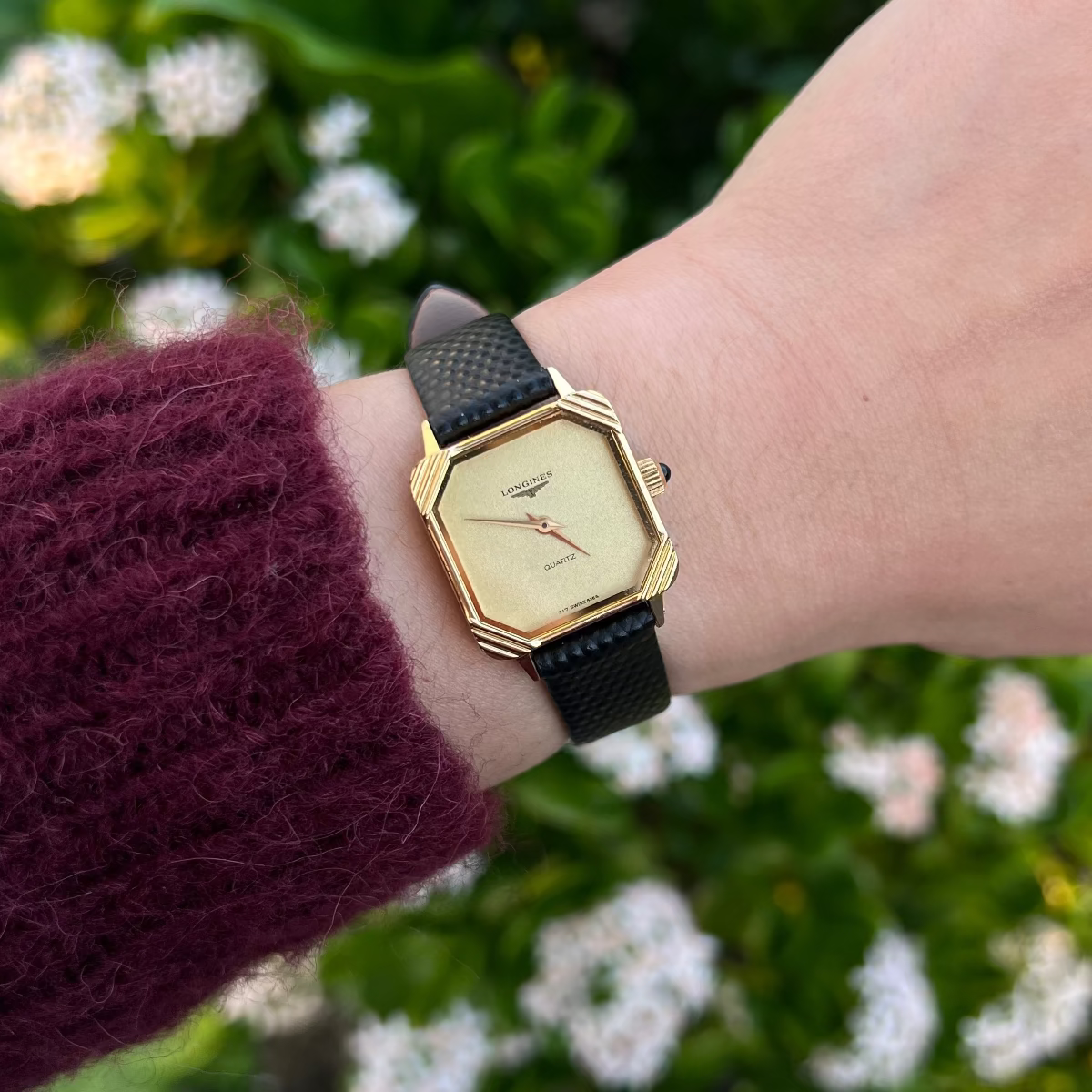 Longines women’s watch with a hexagonal gold dial, black leather strap and blue stone crown worn on the wrist.