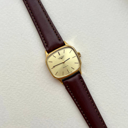 Longines soft octagonal gold-tone ladies watch with pale champagne dial and chocolate-brown leather strap on a textured white background.