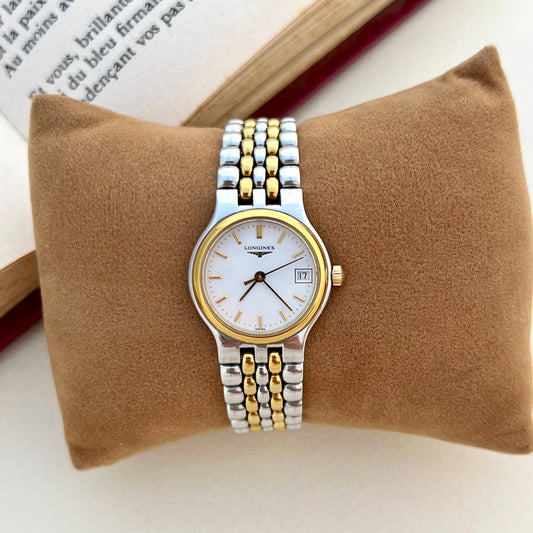 Longines two-tone ladies watch with gold bezel displayed on a cushion beside an open book.
