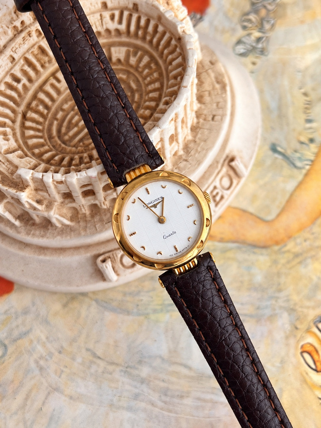 Longines white dial women’s watch with gold tone bezel and brown leather strap displayed beside a sculptural Colosseum model.