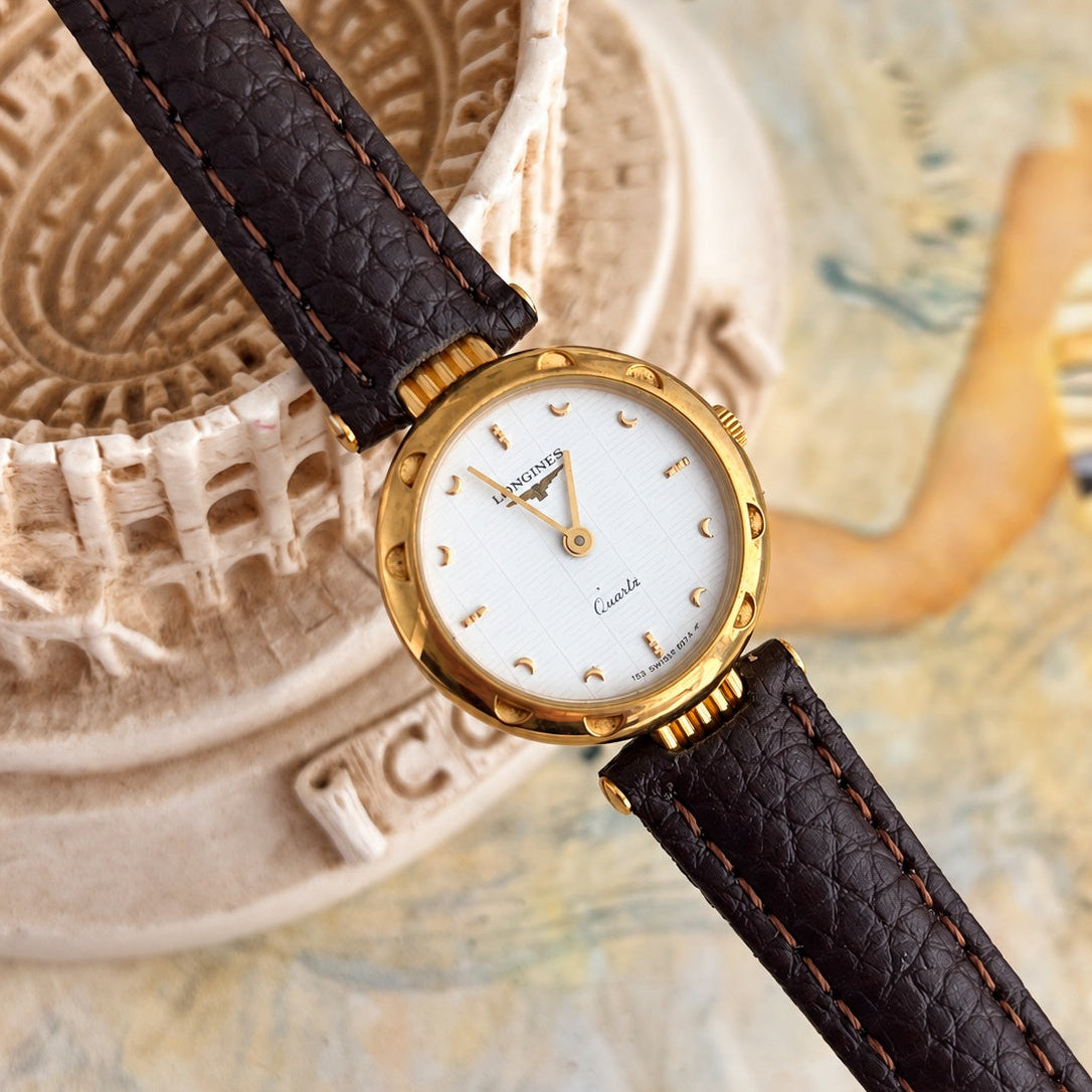 Close angled view showing gold tone crescent hour markers and faint linear pattern on the white Longines dial.