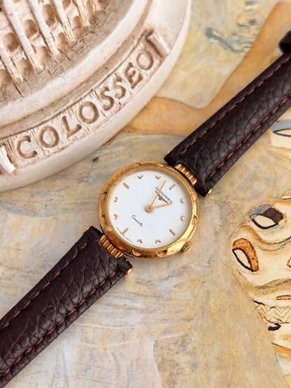 Longines decorated gold tone round bezel, fluted lugs and slender gold tone hands shown against an art print.