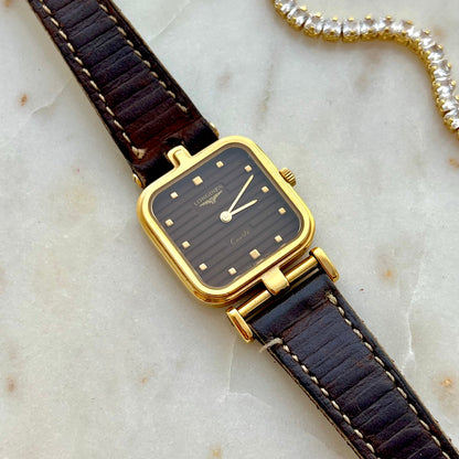 Longines women’s gold tone dress watch with black ribbed dial, square case, gold stud markers and brown lizard grain leather strap styled on marble with crystal jewellery.