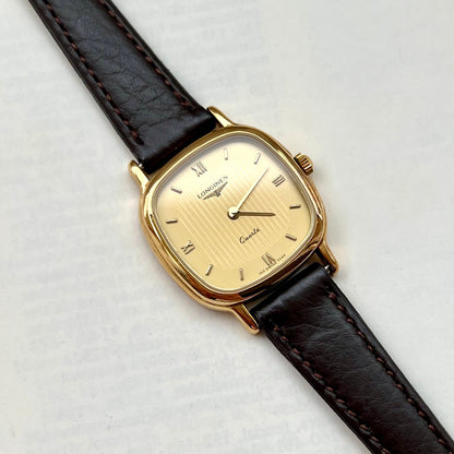 Longines ladies gold cushion watch with champagne striped dial and dark brown leather strap on light background