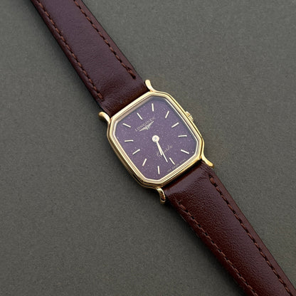 Longines ladies gold watch with deep burgundy dial and brown leather strap on wrist beside diamond tennis bracelet