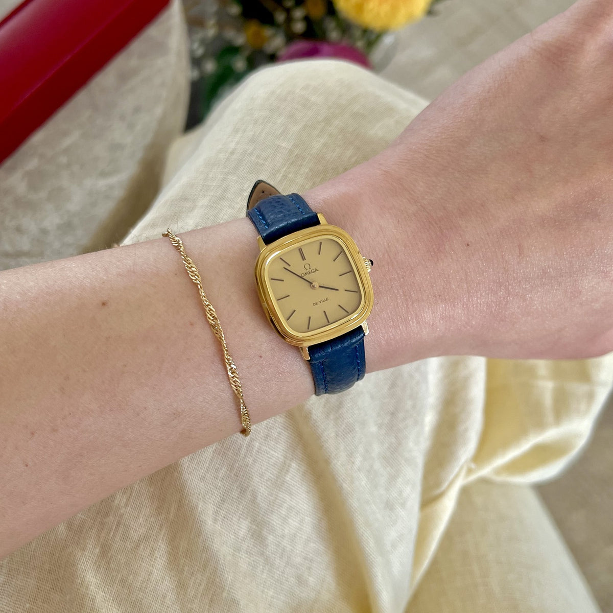 Omega De Ville gold-tone cushion women's watch with champagne dial and navy blue textured strap, worn on wrist