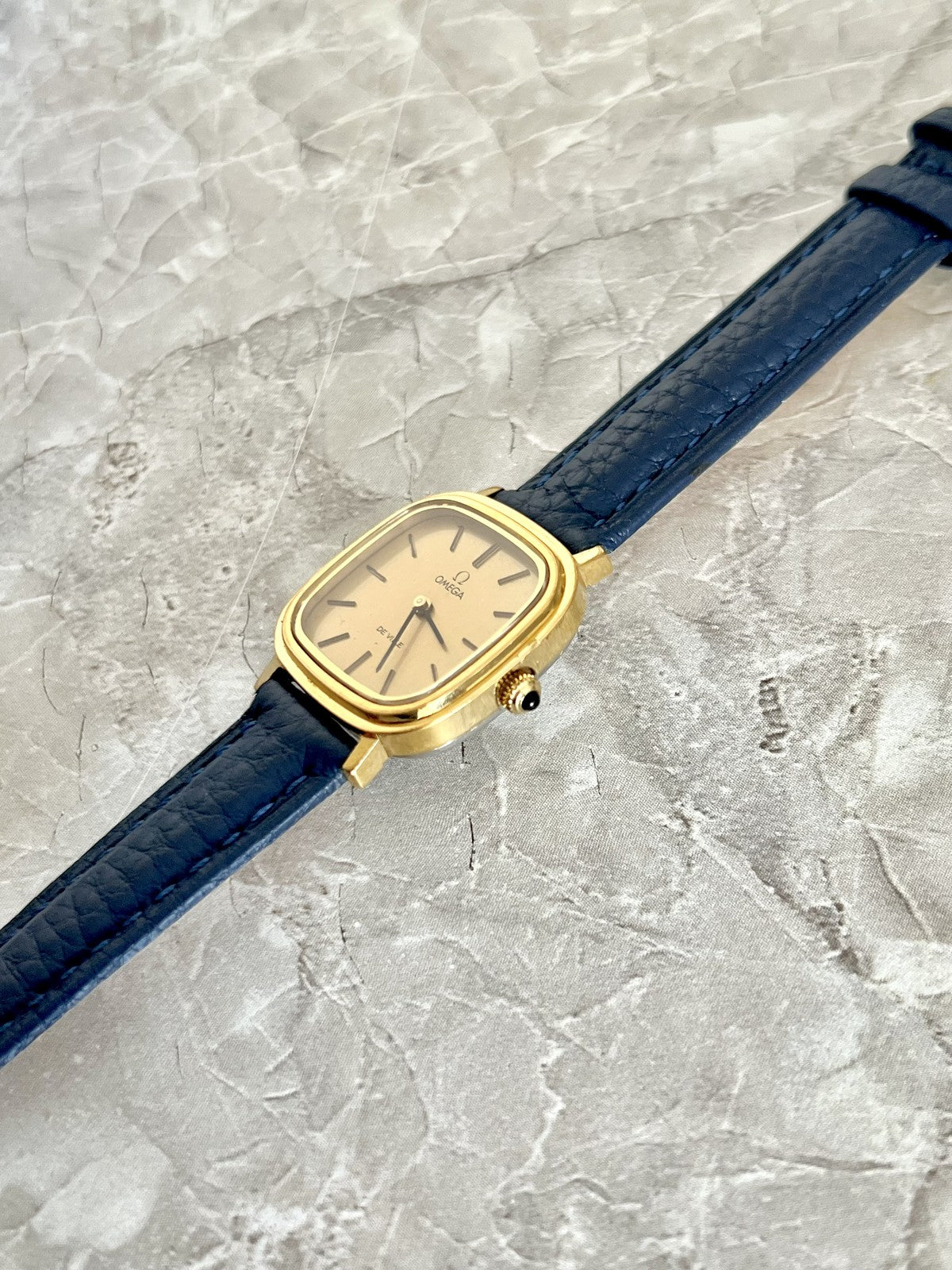 Angled view of Omega De Ville gold-tone cushion case and black-tipped crown on navy blue strap