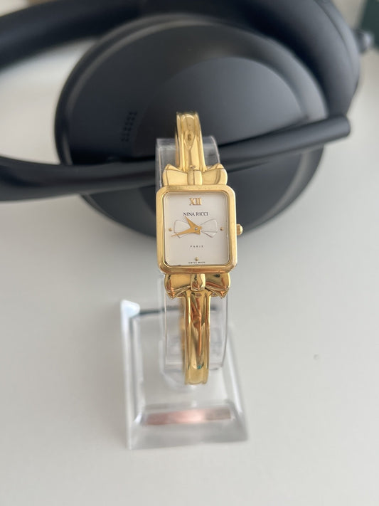 Nina Ricci D950 women’s watch with a rectangular white bow dial and gold plated fixed position bangle, shown front on.