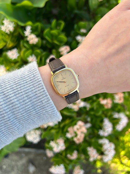 Omega Cushioned Gold Toned Woman's Watch worn on a wrist with a dark leather strap and champagne dial.