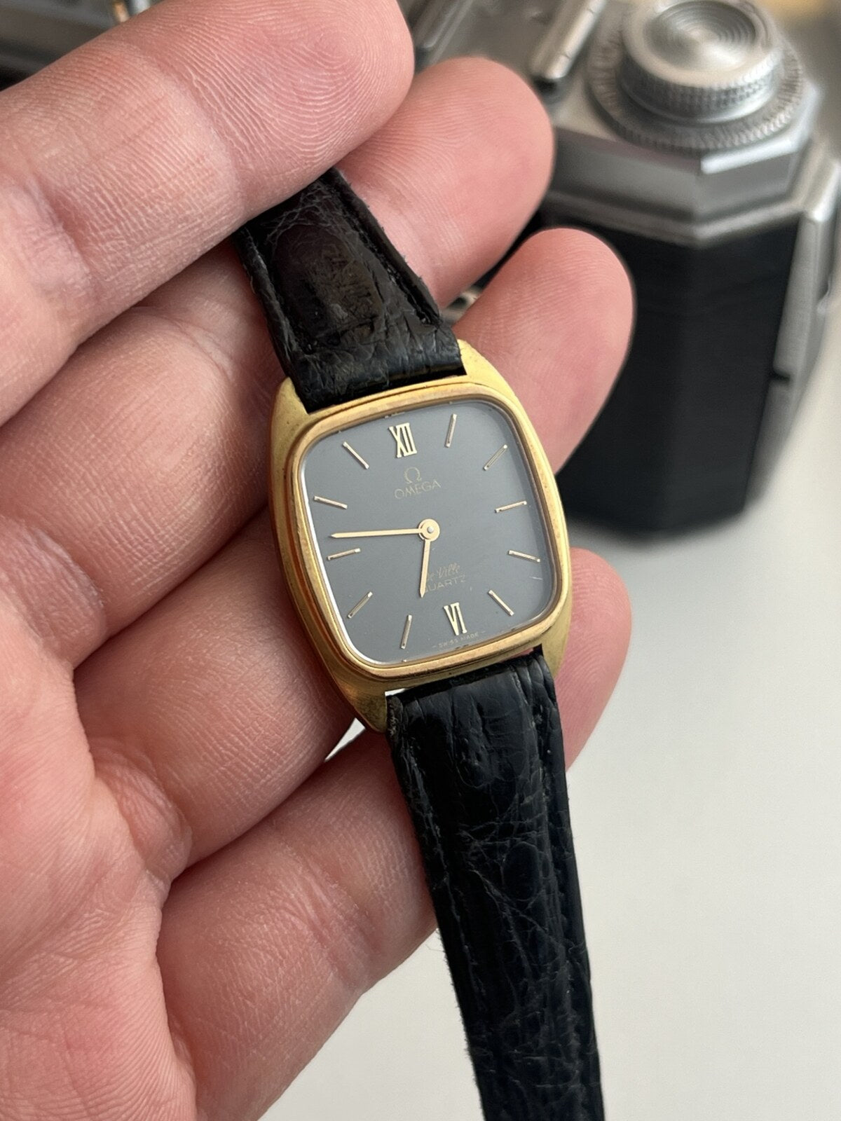 Omega De Ville women’s watch with an ash grey dial, rounded gold tone case and black aftermarket leather strap.