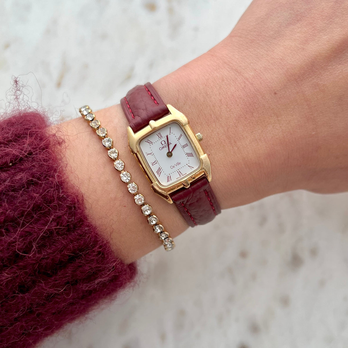 Omega De Ville gold women’s watch with burgundy Roman numerals and burgundy leather strap worn on wrist