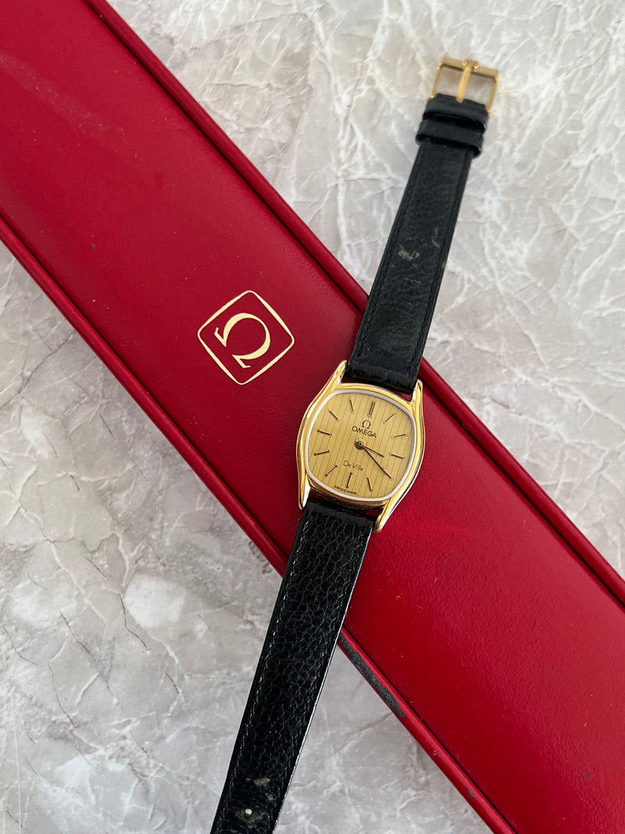 Flat-lay view of Omega De Ville quartz watch with gold-tone cushion case, champagne dial and black leather strap beside a red box