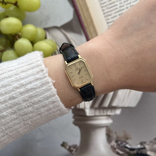 Omega De Ville champagne dial watch with a clipped corner gold tone frame and black patterned leather strap, worn on a 15 cm wrist beside grapes and an open book.