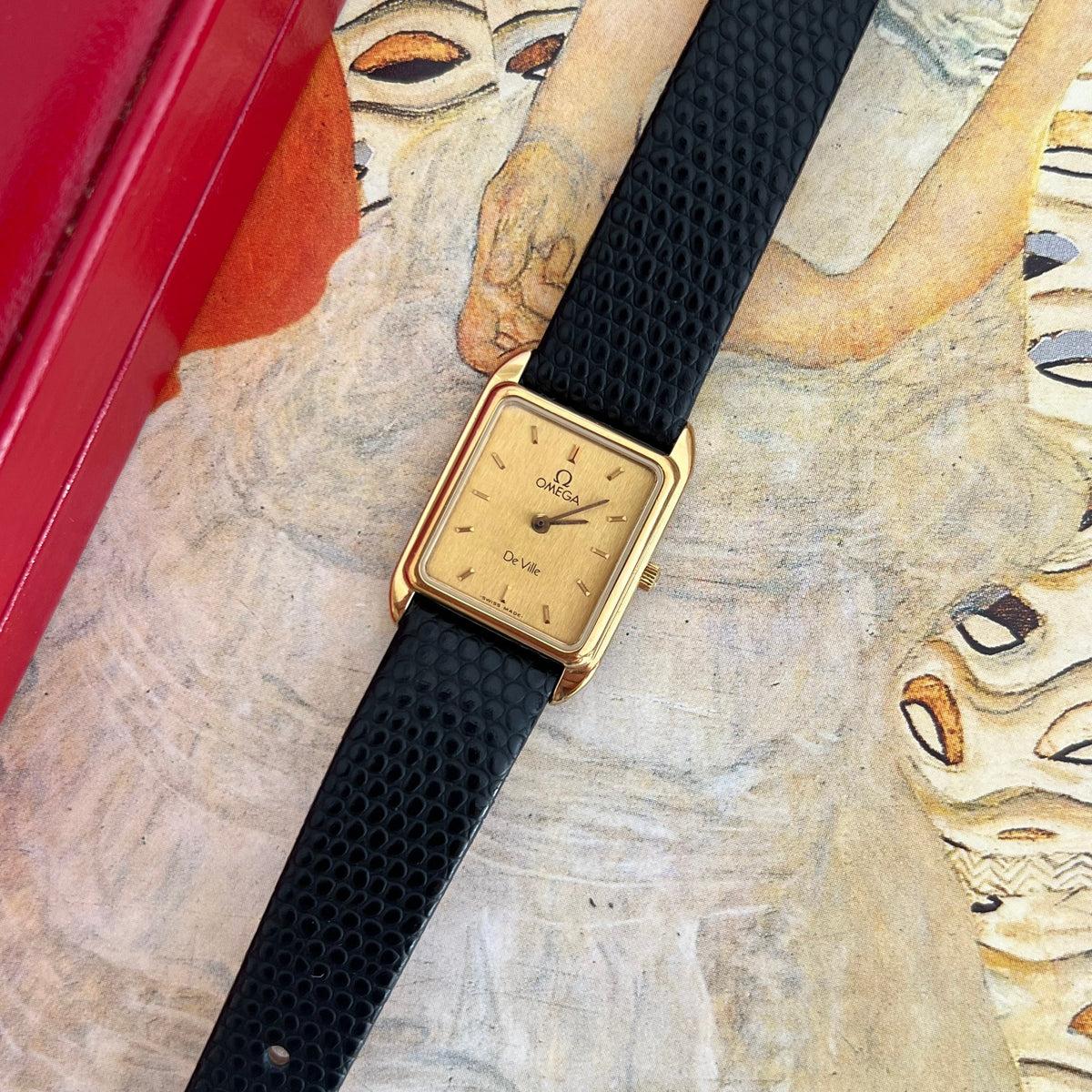 Omega De Ville women's watch with a champagne baton dial, gold tone rectangular case and black textured aftermarket strap.