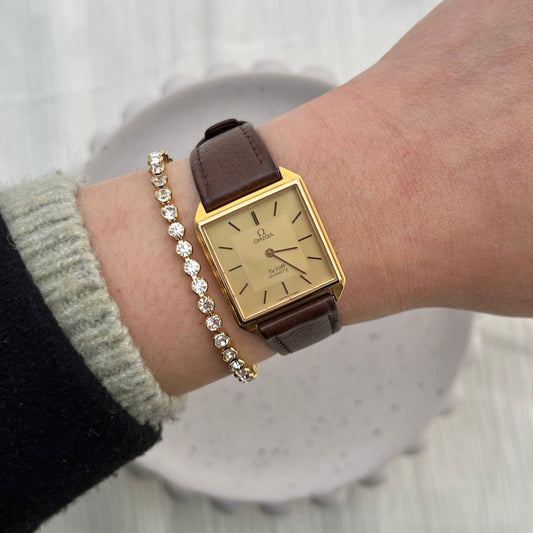Omega De Ville gold-tone women's watch with champagne dial and brown leather strap, shown on a wrist.