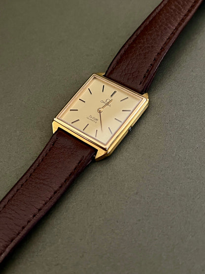 Omega De Ville gold-tone watch with black baton markers and slim hands on a champagne dial.
