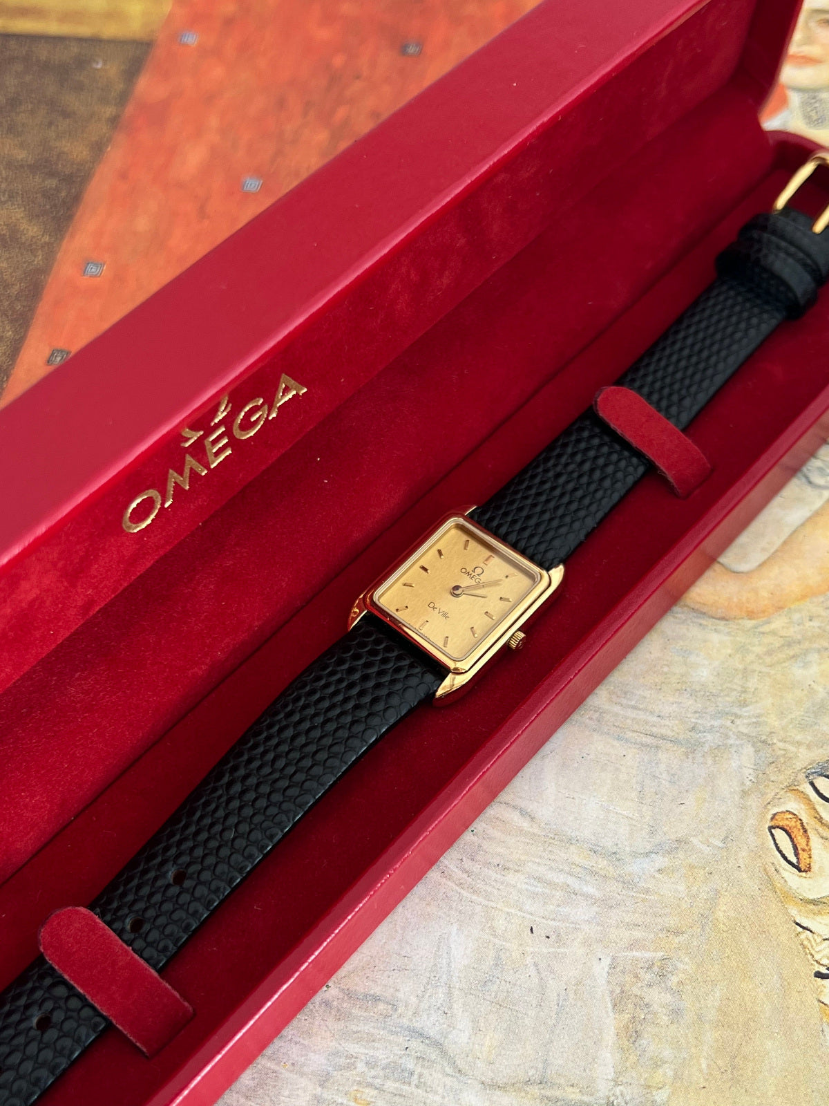 Omega De Ville champagne dial watch presented inside its original red Omega box with the black textured strap visible.