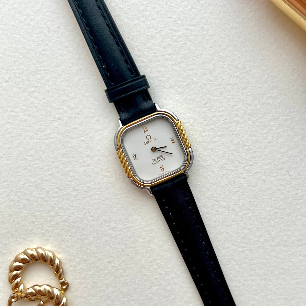 Omega De Ville cushion-case watch with black leather strap and twisted gold column bezel, photographed with red lipstick.