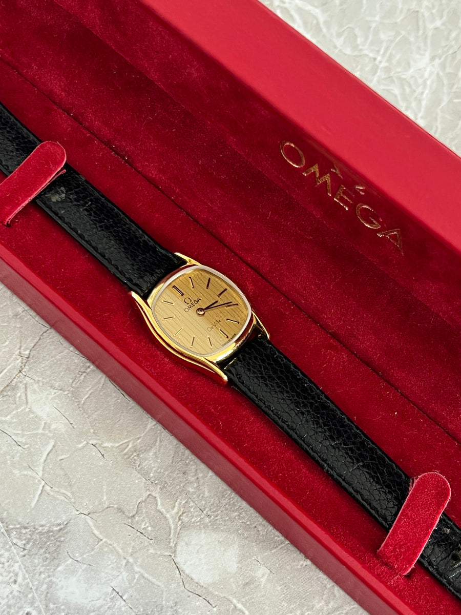 Angled view of Omega De Ville gold-tone cushion watch showing its vertically textured champagne dial and black leather strap