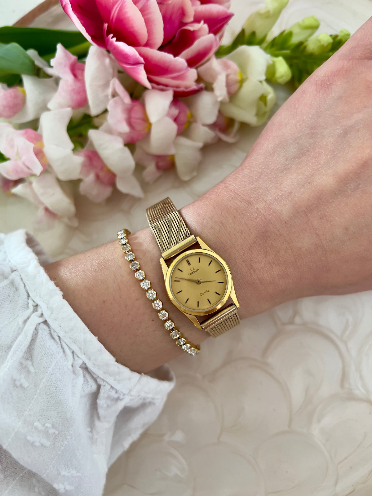 Omega De Ville gold-tone watch with mesh bracelet shown on a wrist.