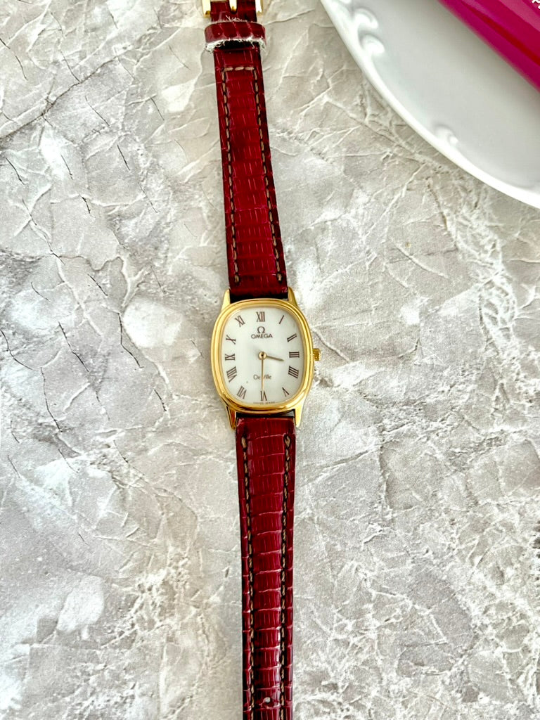 Omega De Ville women’s gold-tone oval watch with white Roman numeral dial and red crocodile-pattern strap, full front view