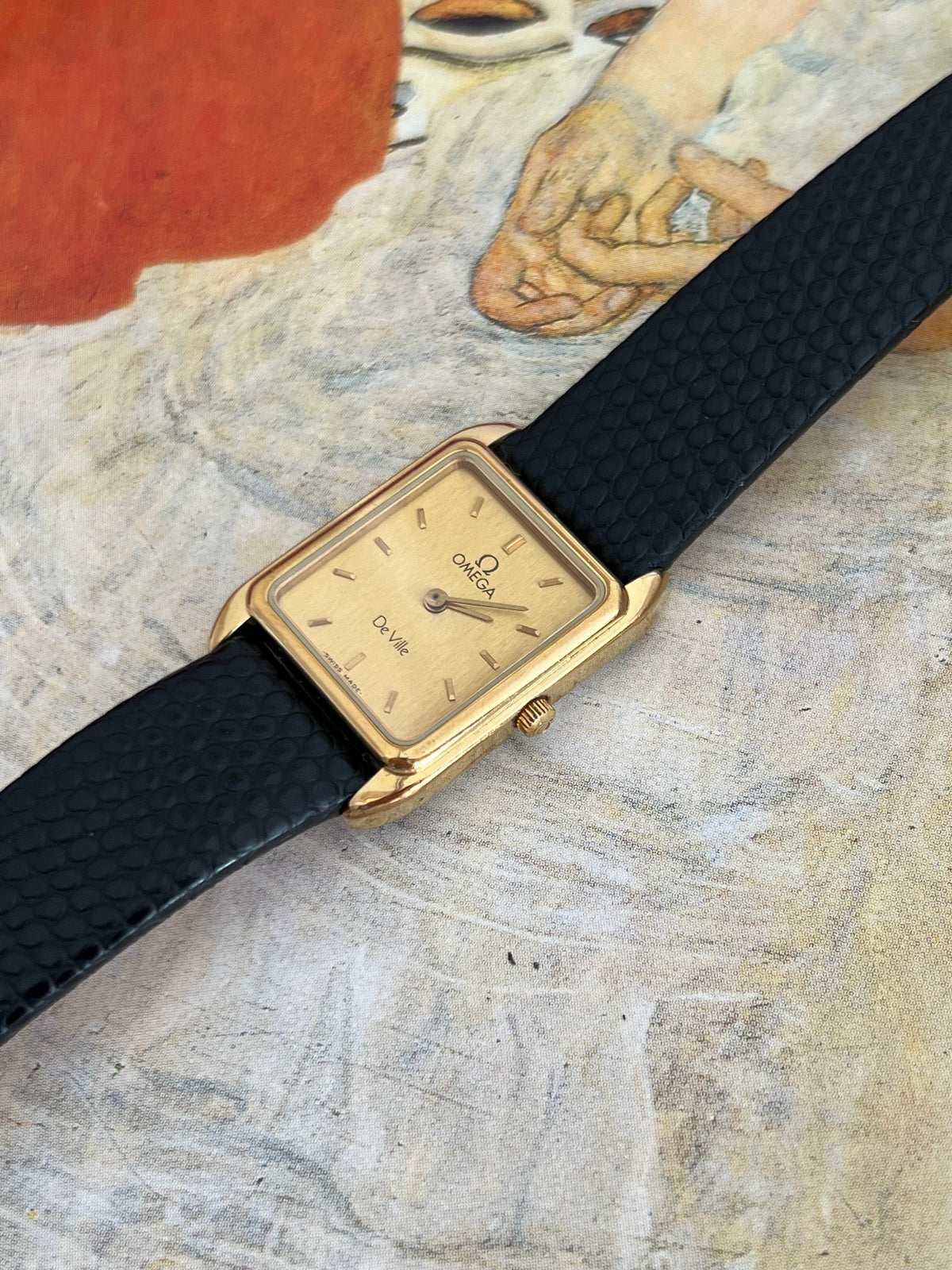 Angled close view of the Omega De Ville gold tone rectangular watch showing the champagne dial, fluted crown and black textured strap.