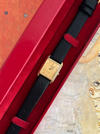 Closer view of the Omega De Ville gold tone rectangular watch and black strap resting in the red Omega presentation box.