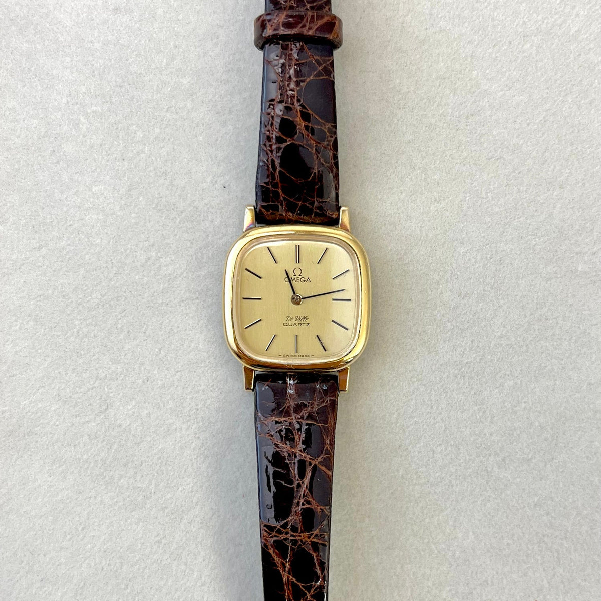Gold Omega De Ville cushion watch with brown leather strap on a light grey background.