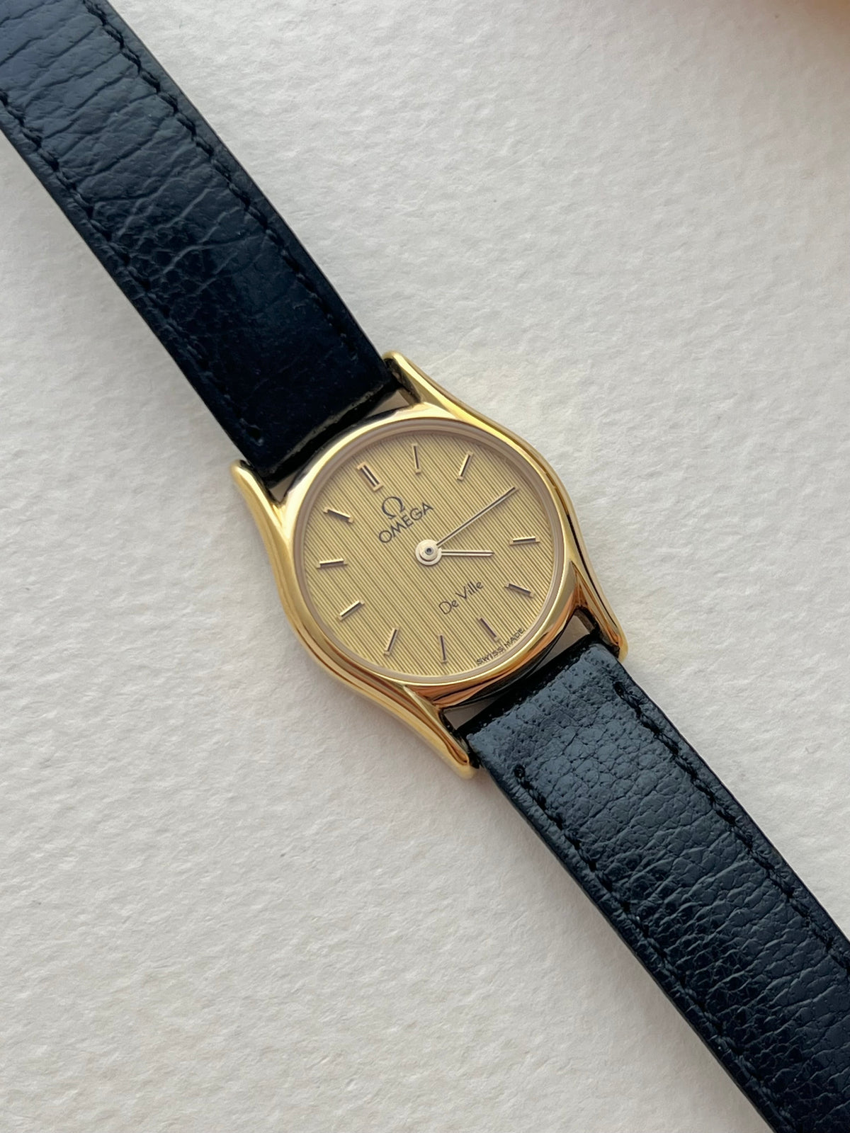 Omega De Ville women's gold-tone watch with vertical patterned dial and black-accented hour markers.