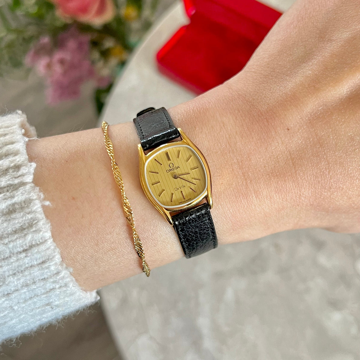 Omega De Ville women's gold-tone cushion watch with textured champagne dial and black leather strap, shown on the wrist