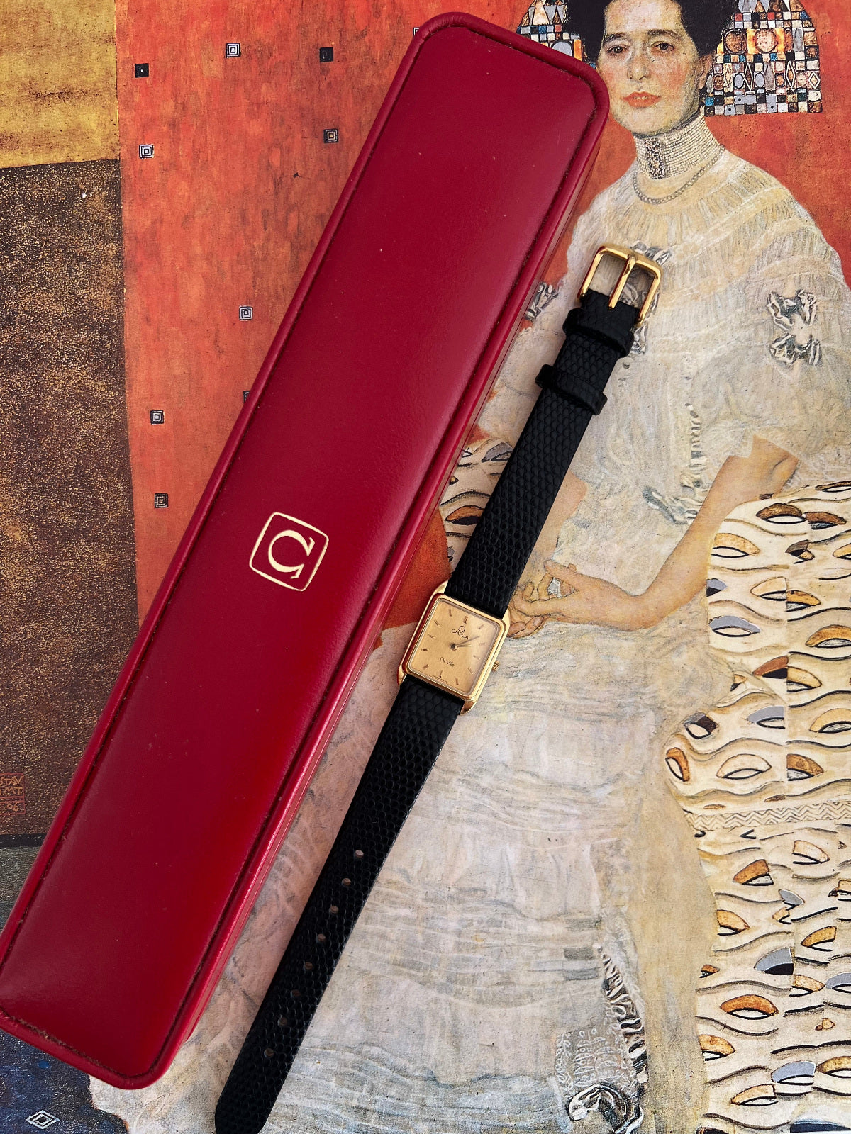 Full-length Omega De Ville women's watch with black aftermarket strap and gold tone pin buckle beside its original red Omega box.