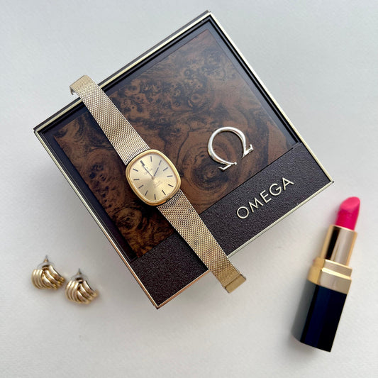 Omega De Ville watch with a champagne gold dial, rounded square case and gold tone mesh bracelet displayed on an Omega box