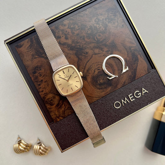 Omega De Ville women’s watch with a champagne dial, softly squared gold tone case and fine mesh bracelet displayed on an Omega box