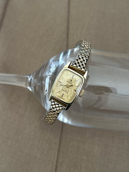 Close view of the gold linen textured dial, baton markers and two hands on the Omega De Ville women’s watch