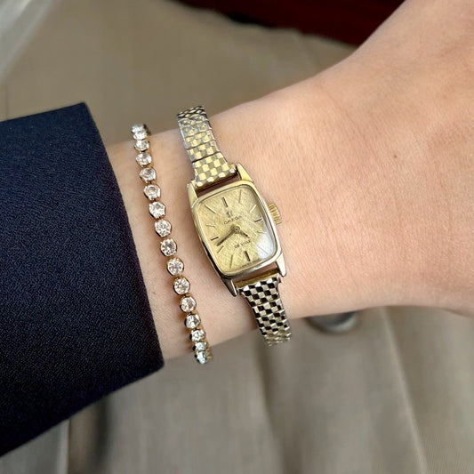 Omega De Ville women’s watch with a gold linen textured dial and two tone elastic bracelet, worn beside a crystal bracelet